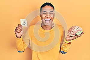 Young african american guy holding smile reminder and brain smiling and laughing hard out loud because funny crazy joke