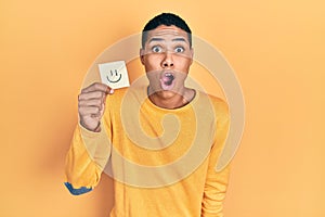 Young african american guy holding smile reminder scared and amazed with open mouth for surprise, disbelief face