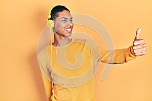 Young african american guy listening to music using headphones looking proud, smiling doing thumbs up gesture to the side