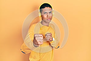 Young african american guy listening to music using headphones ready to fight with fist defense gesture, angry and upset face,