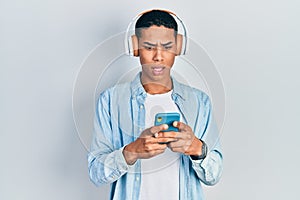 Young african american guy using smartphone wearing headphones scared and amazed with open mouth for surprise, disbelief face