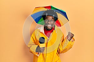 Young african american journalist man wearing yellow raincoat and umbrella cap screaming proud, celebrating victory and success