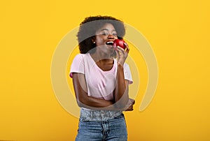 Young african american lady with beautiful smile eating tasty red apple, promoting healthy nutrition, yellow background