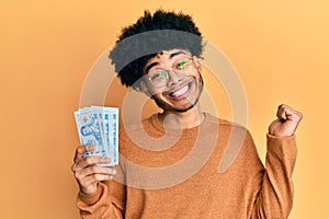 Young african american man with afro hair holding 50 thai baht banknotes screaming proud, celebrating victory and success very