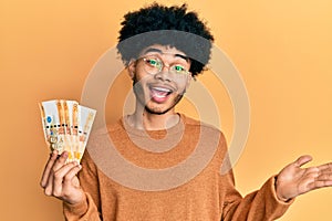 Young african american man with afro hair holding 500 philippine peso banknotes celebrating achievement with happy smile and