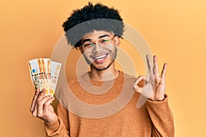 Young african american man with afro hair holding 500 philippine peso banknotes doing ok sign with fingers, smiling friendly