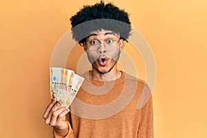 Young african american man with afro hair holding 500 philippine peso banknotes scared and amazed with open mouth for surprise,