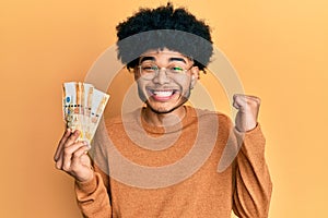 Young african american man with afro hair holding 500 philippine peso banknotes screaming proud, celebrating victory and success