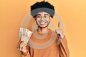 Young african american man with afro hair holding 500 philippine peso banknotes smiling with an idea or question pointing finger