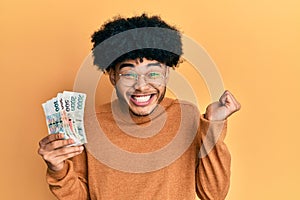 Young african american man with afro hair holding czech koruna banknotes screaming proud, celebrating victory and success very