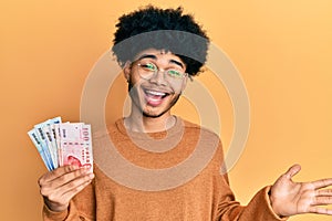 Young african american man with afro hair holding new taiwan dollars banknotes celebrating achievement with happy smile and winner