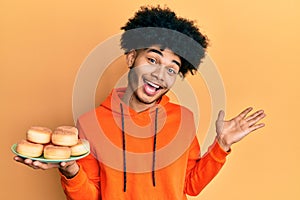 Young african american man with afro hair holding pile of tasty sugar doughnuts celebrating achievement with happy smile and