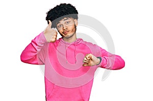 Young african american man with afro hair wearing casual pink sweatshirt doing thumbs up and down, disagreement and agreement