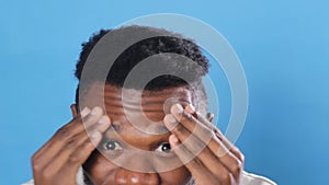 a young African American man attentively examines his face looking into the camera close-up blue background.