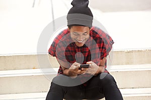 Young african american man with beanie sitting on steps outside and looking at mobile phone