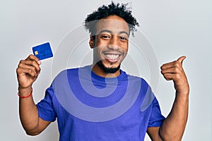 Young african american man with beard holding credit card pointing thumb up to the side smiling happy with open mouth