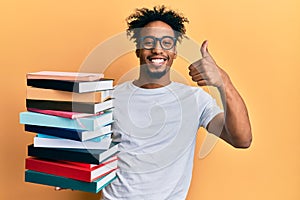Young african american man with beard holding a pile of books smiling happy and positive, thumb up doing excellent and approval