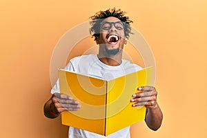 Young african american man with beard reading a book wearing glasses smiling and laughing hard out loud because funny crazy joke