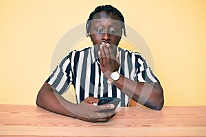 Young african american man with braids using smartphone sitting on the table covering mouth with hand, shocked and afraid for