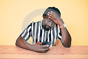 Young african american man with braids using smartphone sitting on the table stressed and frustrated with hand on head, surprised