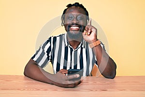 Young african american man with braids using smartphone sitting on the table surprised with an idea or question pointing finger