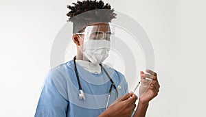 Young African American man doctor using syringe to suck vaccines or liquid medicine out of vial bottle with mask and face shield