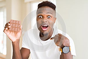 Young african american man eating chocolate bar with surprise face pointing finger to himself