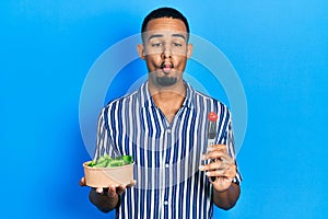 Young african american man eating salad making fish face with mouth and squinting eyes, crazy and comical