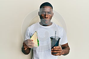 Young african american man eating sandwich and drinking soda making fish face with mouth and squinting eyes, crazy and comical