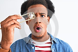 Young african american man eating sushi using chopsticks over isolated white background scared in shock with a surprise face,