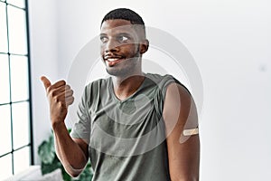 Young african american man getting vaccine showing arm with band aid pointing thumb up to the side smiling happy with open mouth