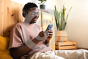Young african american man with glasses relaxing at home listening to music with headphones while using mobile phone.