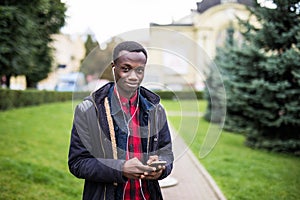 Young African American man in headphone walking at sunny city and enjoying to listen to music on his smart phone.