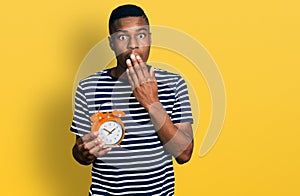 Young african american man holding alarm clock covering mouth with hand, shocked and afraid for mistake