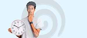 Young african american man holding big clock covering mouth with hand, shocked and afraid for mistake