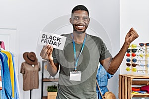 Young african american man holding black friday banner at retail shop screaming proud, celebrating victory and success very