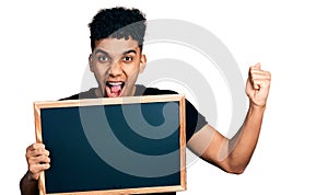 Young african american man holding blackboard screaming proud, celebrating victory and success very excited with raised arms