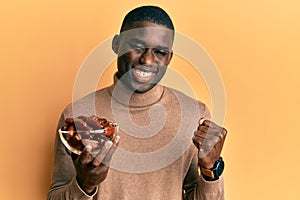 Young african american man holding bowl with dates screaming proud, celebrating victory and success very excited with raised arm