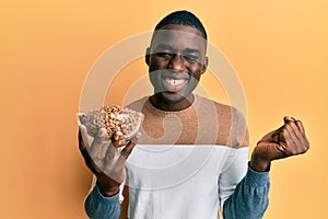 Young african american man holding bowl with lentils screaming proud, celebrating victory and success very excited with raised arm