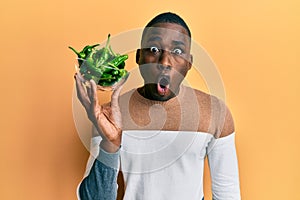 Young african american man holding bowl with small green peppers scared and amazed with open mouth for surprise, disbelief face