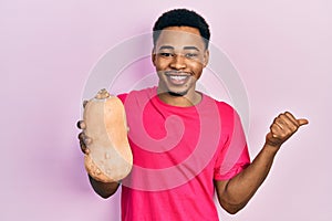 Young african american man holding healthy fresh pumpkin pointing thumb up to the side smiling happy with open mouth