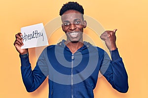 Young african american man holding healthy hashtag paper pointing thumb up to the side smiling happy with open mouth