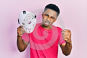 Young african american man holding hockey mask annoyed and frustrated shouting with anger, yelling crazy with anger and hand