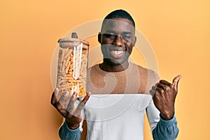 Young african american man holding jar of macaroni pasta pointing thumb up to the side smiling happy with open mouth