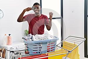 Young african american man holding magnifying glass looking for stain at clothes screaming proud, celebrating victory and success