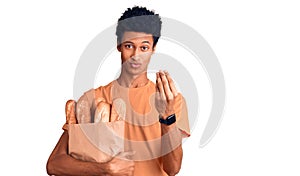 Young african american man holding paper bag with bread doing money gesture with hands, asking for salary payment, millionaire