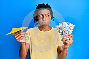 Young african american man holding paper plane and dollars puffing cheeks with funny face