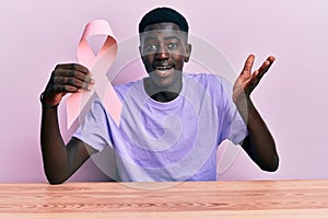 Young african american man holding pink cancer ribbon sitting on the table celebrating achievement with happy smile and winner