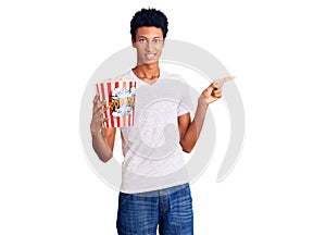 Young african american man holding popcorn smiling happy pointing with hand and finger to the side