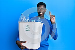 Young african american man holding recycling wastebasket with plastic bottles surprised with an idea or question pointing finger